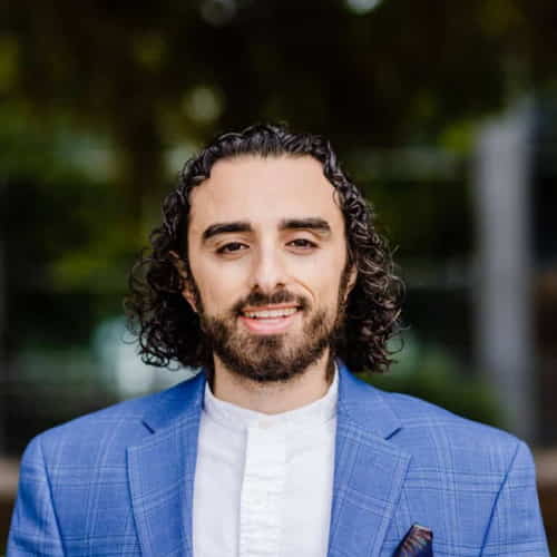 A person with curly hair and a beard is smiling, wearing a light blue blazer and white shirt, embodying the professionalism sought to hire top-tier talent in Eastern Europe. The background is blurred, with hints of green foliage.